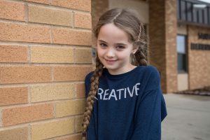 An elementary girl in a blue shirt against a brick wall