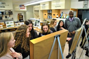 In a classroom, students and adults look at and discuss paintings propped up on easels.