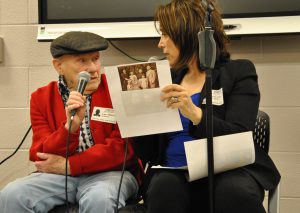 Man and woman sitting, holding microphones, seaking to each other. Woman holds a sheet of paper with an old photo of a family.