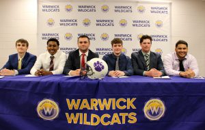 Six high school students wearing suits and ties sit at a table that is covered with a purple banner with gold writing. The banner says Warwick Wildcats.