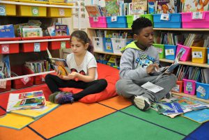 Two students sitting on a classroom rugs select books stacked in bins in their classroom library.s