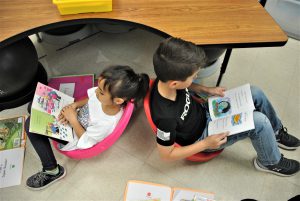 Two students sit back to back in scoop chairs reading picture books.