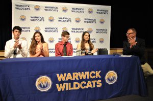Four students sitting and athletics director sitting and applauding at a draped table with district logos. A backdrop is imprinted with Wildcats and district logos.
