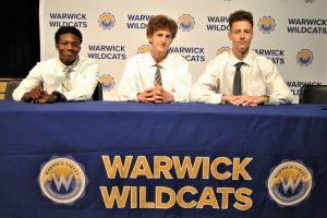 Three students sitting at draped table with district logos. A backdrop is imprinted with Wildcats and district logos.