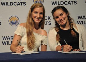 Two students sitting at draped table, signing National Letter of Intent.. A backdrop is imprinted with Wildcats and district logos
