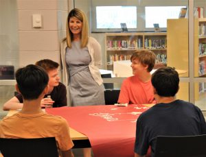 Four students sit at a table engage in conversation with an educator standing by. The setting it the school's media center. aged in conversation