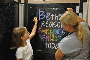 Student and adult hang poster on bathroom door. Poster reads, "Be the reason someone smiles today."
