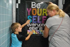 Student and adult set poster on bathroom door. Poster reads, "Be yourself. Everyone else is taken."