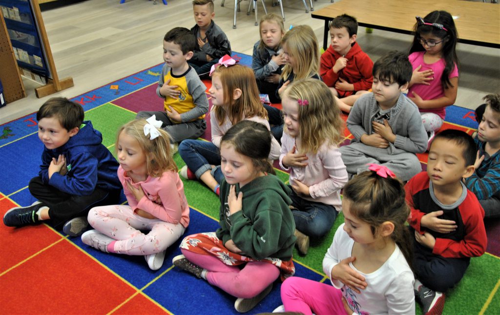 Kindergartners sit on classroom rug, eyes closed, hand over their chest, meditating.