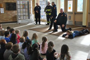 A group of firefighters addresses a student assembly in the school lobby. One firefighter is demonstrating "stop, drop & roll" exercises.
