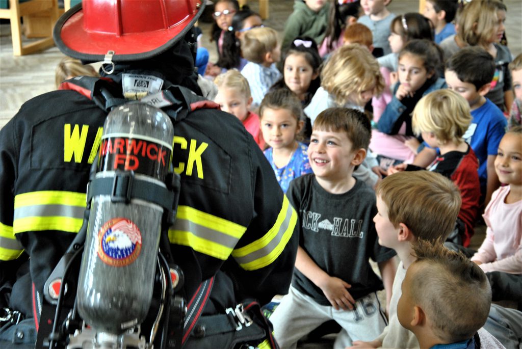 Students gather around a fully-outfitted firefighter who is demonstrating a fire rescue