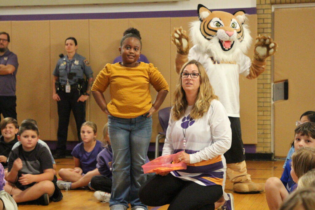 A student answers Jeopardy questions with a teacher next to her and the Wildcat mascot behind her