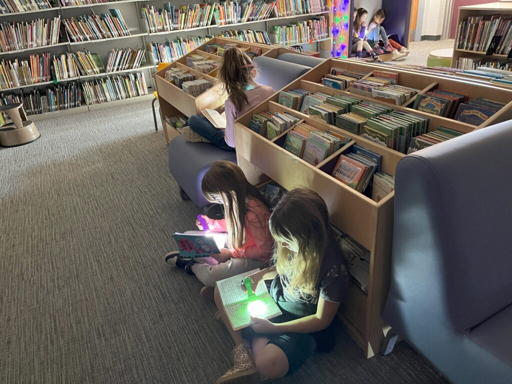 2 students read by flashlight as they sit on the floor up against a bookcase in the library
