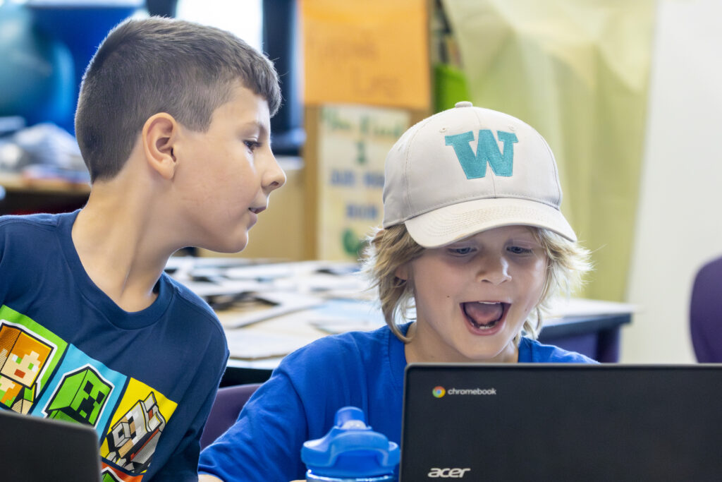 A summer enrichment program student smiles while looking at his laptop while another student looks on