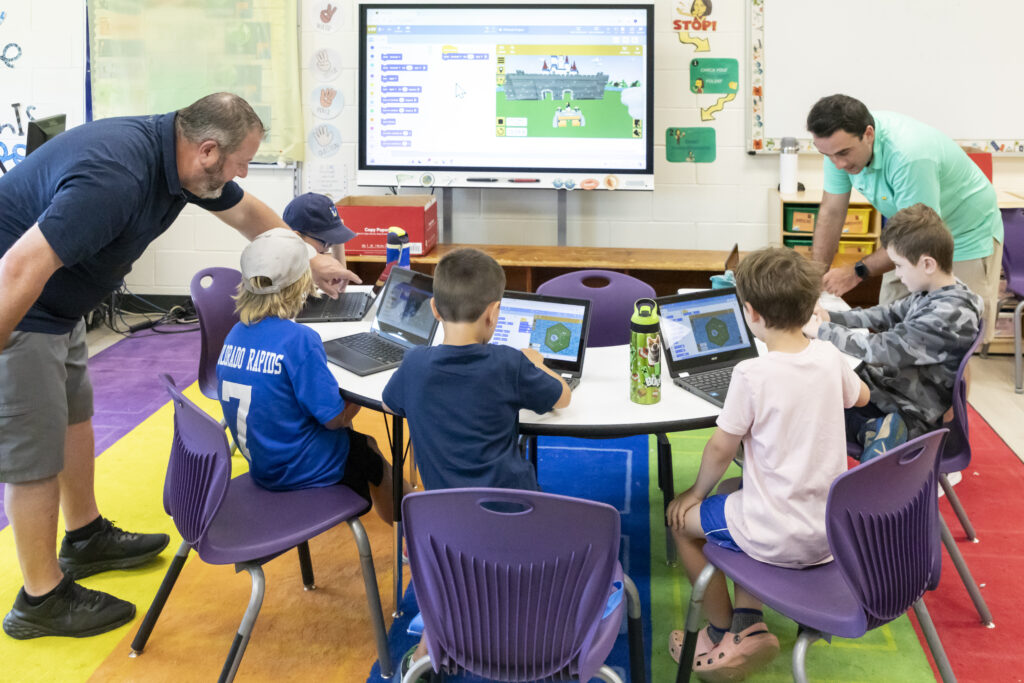 Students in chairs at a table with laptop computers learn coding